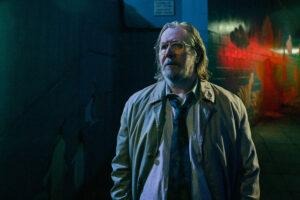 Gary Oldman steht in einer dunklen Straße. Er hat lange haare und sieht etwas fertig aus.
