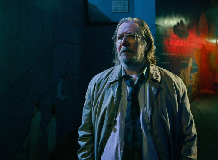 Gary Oldman steht in einer dunklen Straße. Er hat lange haare und sieht etwas fertig aus.